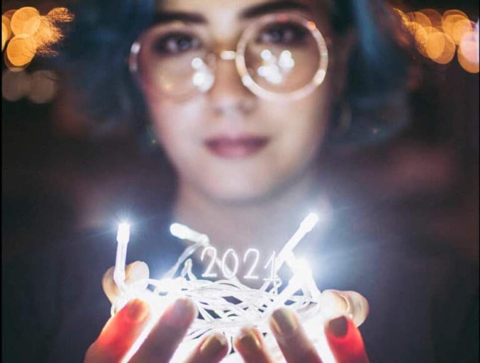 Photographie d'une femme aux cheveux bleus tenant une guirlande de lumières dans ses mains, surmontée des chiffres "2021"